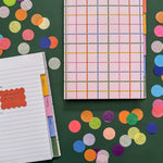 A colorful hardcover notebook with give colorful tabbed sections sits on a dark green surface beside an open tabbed notebook with lined pages. The scene is styled with large round confetti pieces in multiple colors.