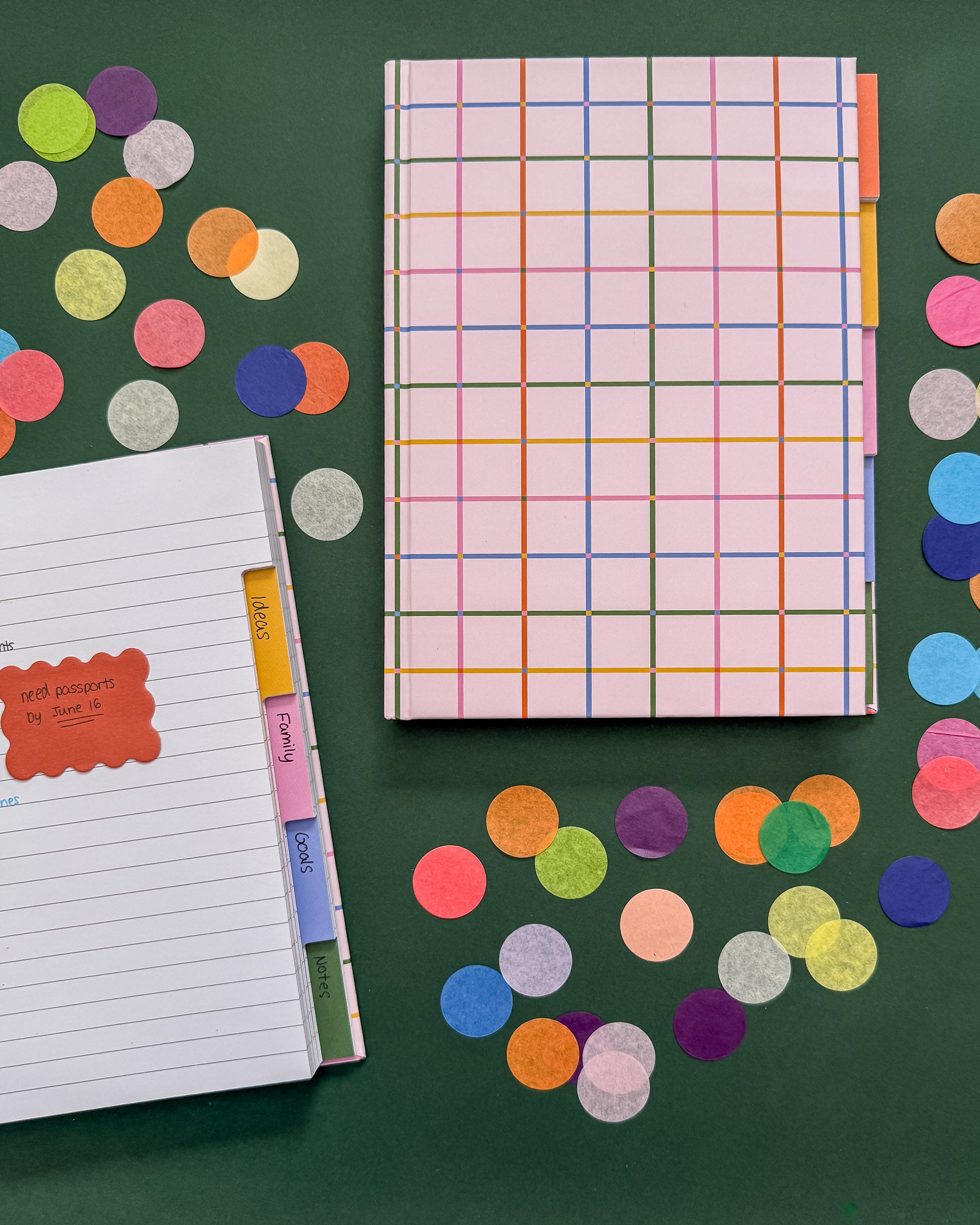 A colorful hardcover notebook with give colorful tabbed sections sits on a dark green surface beside an open tabbed notebook with lined pages. The scene is styled with large round confetti pieces in multiple colors.