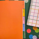 An open notebook with five colorful tabbed sections labeled Ideas, Family, Goods, Motors on a dark green background decorated with large confetti pieces. A closed hardcover notebook with a multicolor plaid pattern is partially visible on the right.
