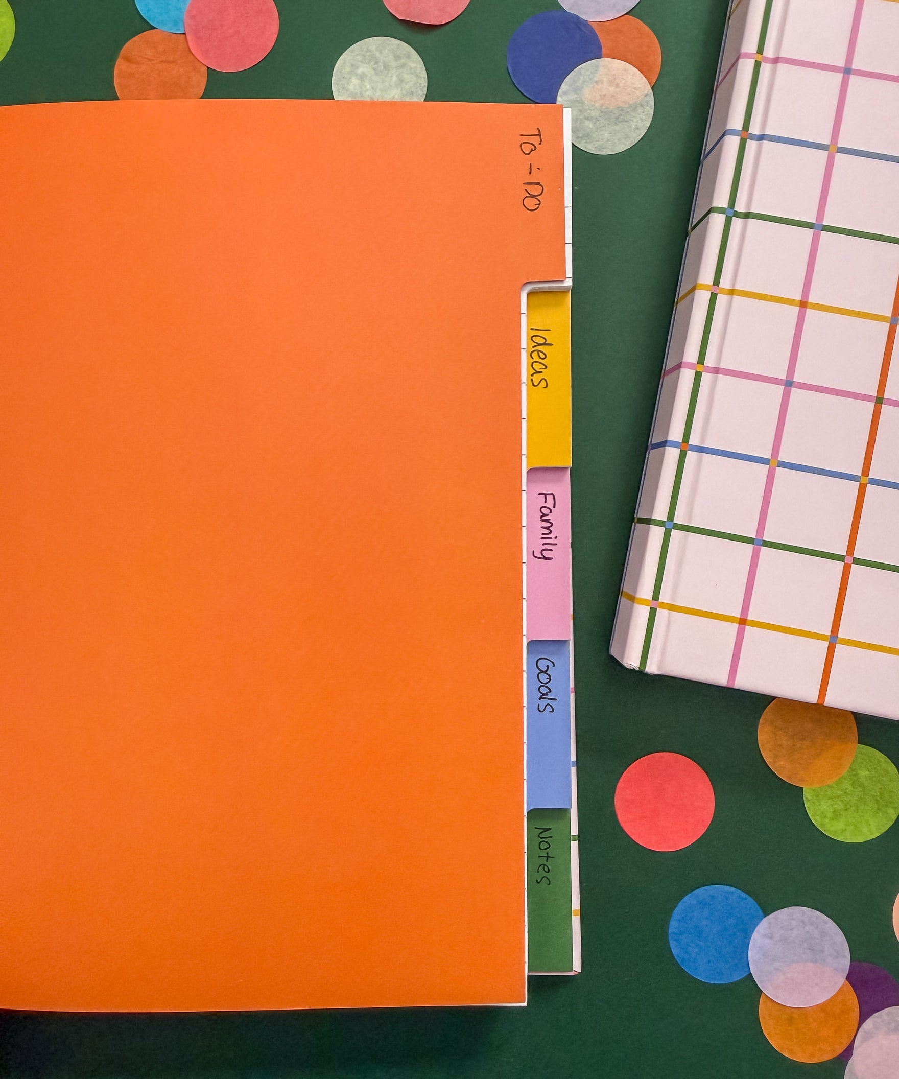 An open notebook with five colorful tabbed sections labeled Ideas, Family, Goods, Motors on a dark green background decorated with large confetti pieces. A closed hardcover notebook with a multicolor plaid pattern is partially visible on the right.