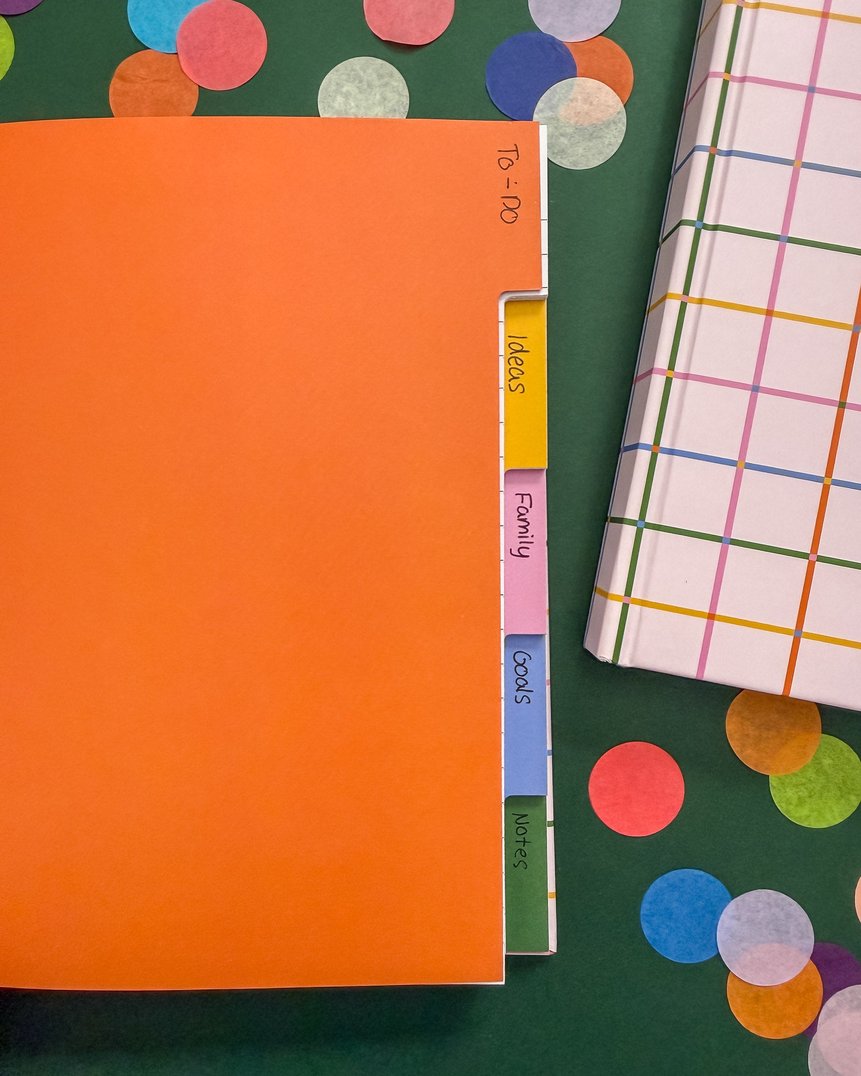 An open notebook with five colorful tabbed sections labeled Ideas, Family, Goods, Motors on a dark green background decorated with large confetti pieces. A closed hardcover notebook with a multicolor plaid pattern is partially visible on the right.