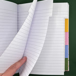 An open lined notebook with a hand lifting two pages, showing white paper with gray horizontal lines and colorful tab dividers on the right side, set against a dark green background.