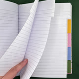 An open lined notebook with a hand lifting two pages, showing white paper with gray horizontal lines and colorful tab dividers on the right side, set against a dark green background.