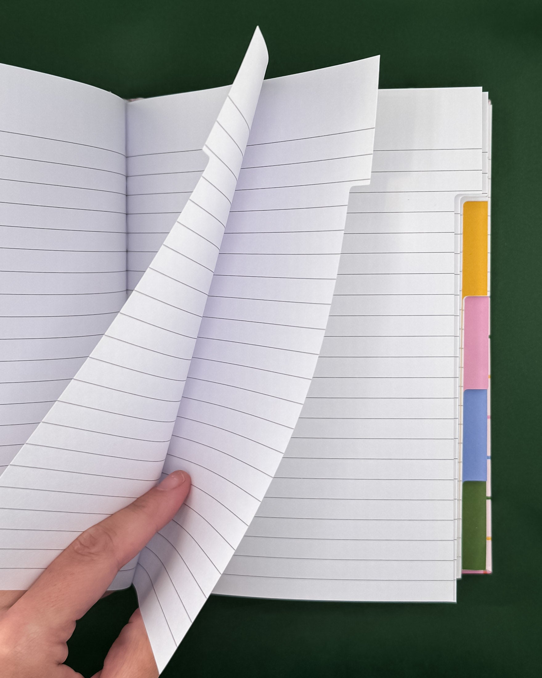 An open lined notebook with a hand lifting two pages, showing white paper with gray horizontal lines and colorful tab dividers on the right side, set against a dark green background.