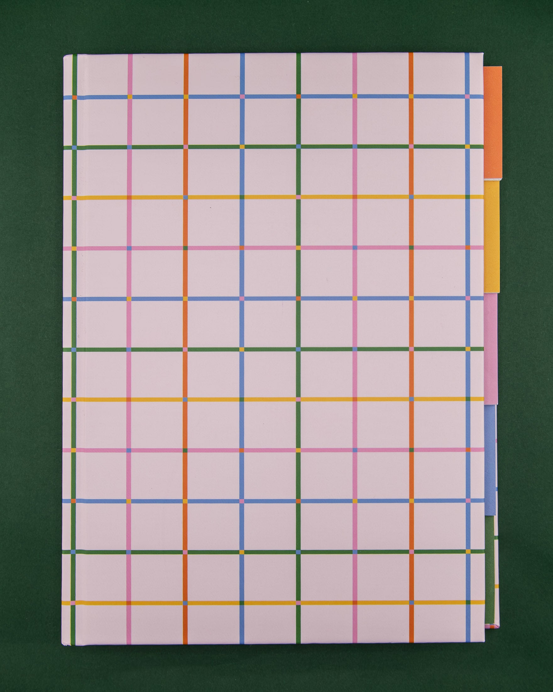 A hardcover notebook with a pale pink cover featuring a colorful plaid pattern and five colorful tabbed sections against a dark green background.