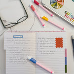 A styled flat-lay of a a lined tabbed notebook on a marble surface surrounded by colorful markers, eyeglasses, and a laptop.