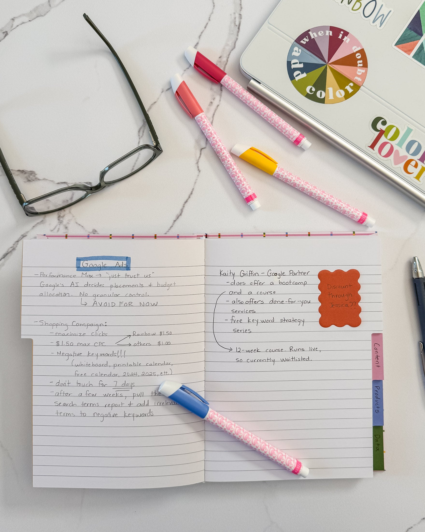 A styled flat-lay of a a lined tabbed notebook on a marble surface surrounded by colorful markers, eyeglasses, and a laptop.