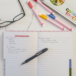 An open notebook with lined pages handwritten notes with a black pen resting on the page, styled on a white marble surface with colorful highlighters, eyeglasses, and a laptop nearby.