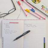 An open notebook with lined pages handwritten notes with a black pen resting on the page, styled on a white marble surface with colorful highlighters, eyeglasses, and a laptop nearby.
