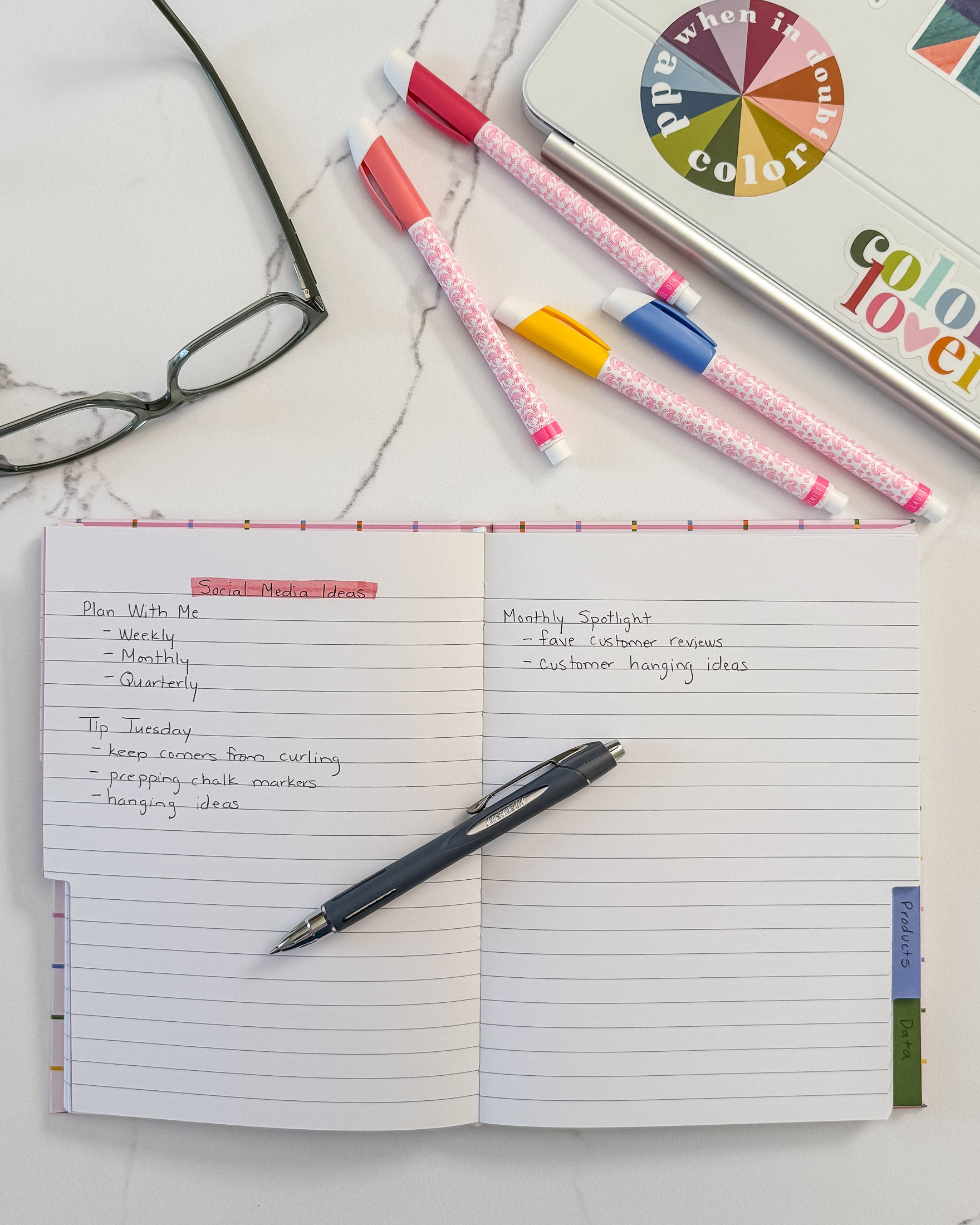 An open notebook with lined pages handwritten notes with a black pen resting on the page, styled on a white marble surface with colorful highlighters, eyeglasses, and a laptop nearby.