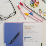 An open notebook with tabbed divided sections and lined pages lying on a white marble surface, styled with a black pen, colorful markers, a pair of eyeglasses, and laptop covered with colorful stickers in the background.