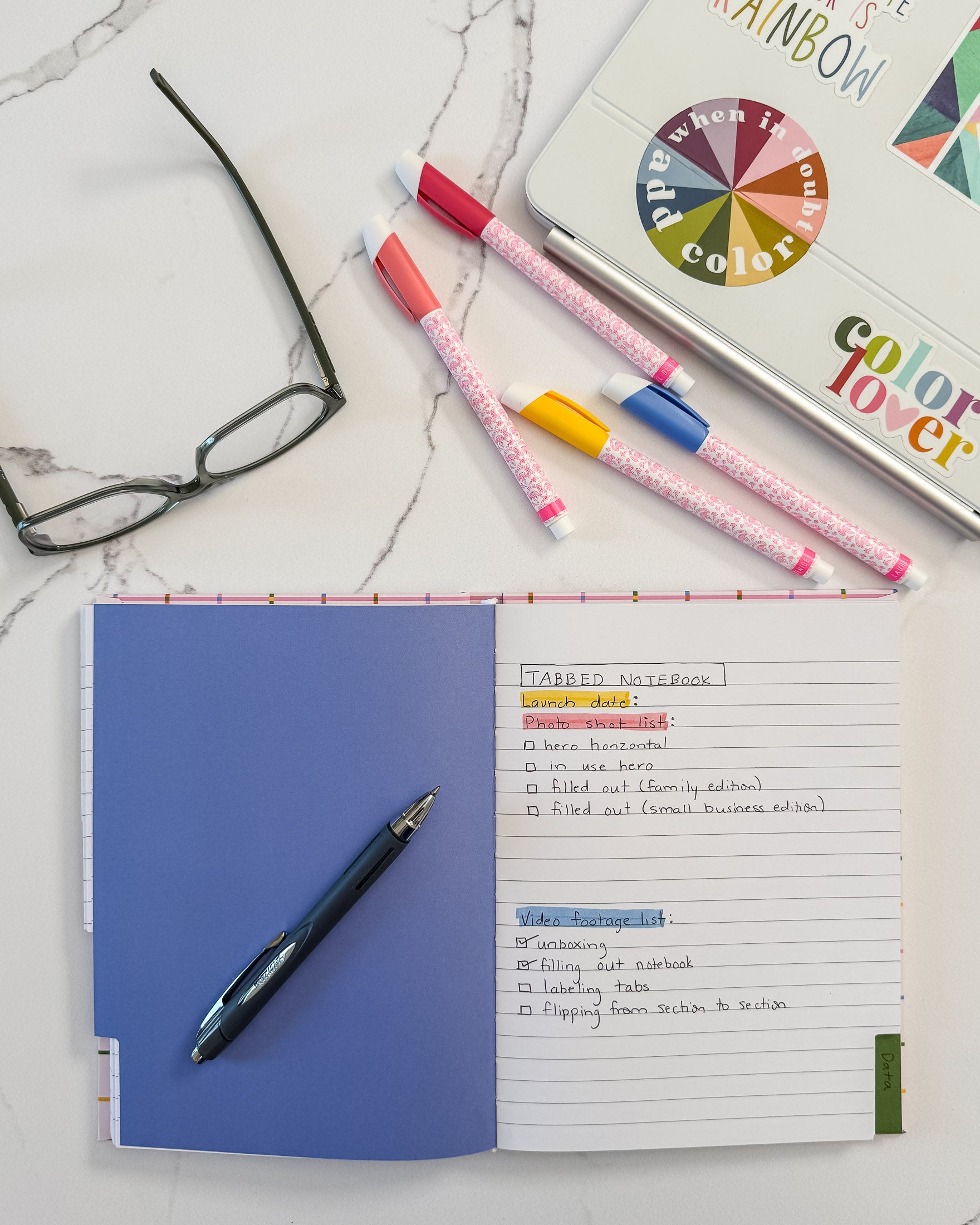 An open notebook with tabbed divided sections and lined pages lying on a white marble surface, styled with a black pen, colorful markers, a pair of eyeglasses, and laptop covered with colorful stickers in the background.
