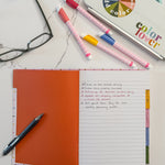 A flat-lay of featuring an open lined hardcover notebook with five colorful tabbed sections, several markers, a blue pen, a pair of eyeglasses, and a laptop with a colorful 'color lover' sticker on a white marble surface.