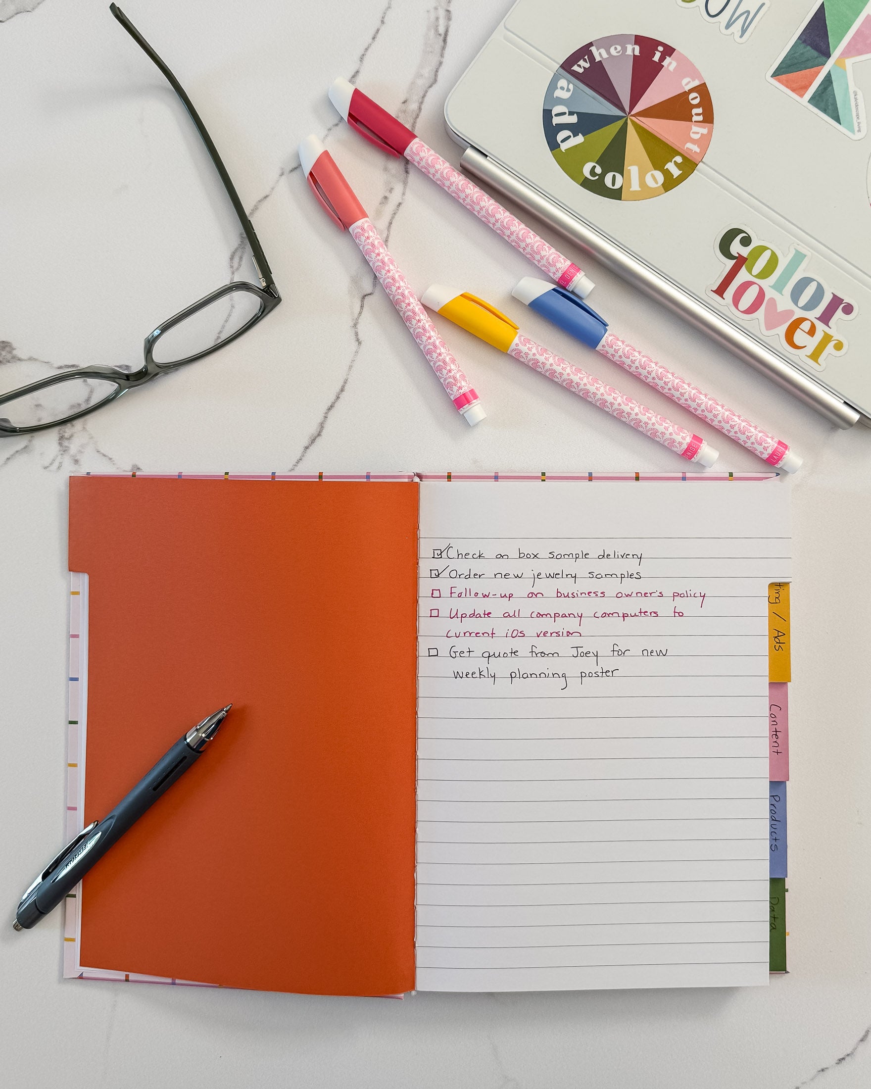A flat-lay of featuring an open lined hardcover notebook with five colorful tabbed sections, several markers, a blue pen, a pair of eyeglasses, and a laptop with a colorful 'color lover' sticker on a white marble surface.