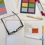 A styled flat-lay of stationery items on a white marble surface, including an open lined notepad with a checklist, a pair of tortoiseshell glasses, colorful scalloped sticky notes, pastel markers, a gold pen, and a notepad from Kaleidoscope Living.