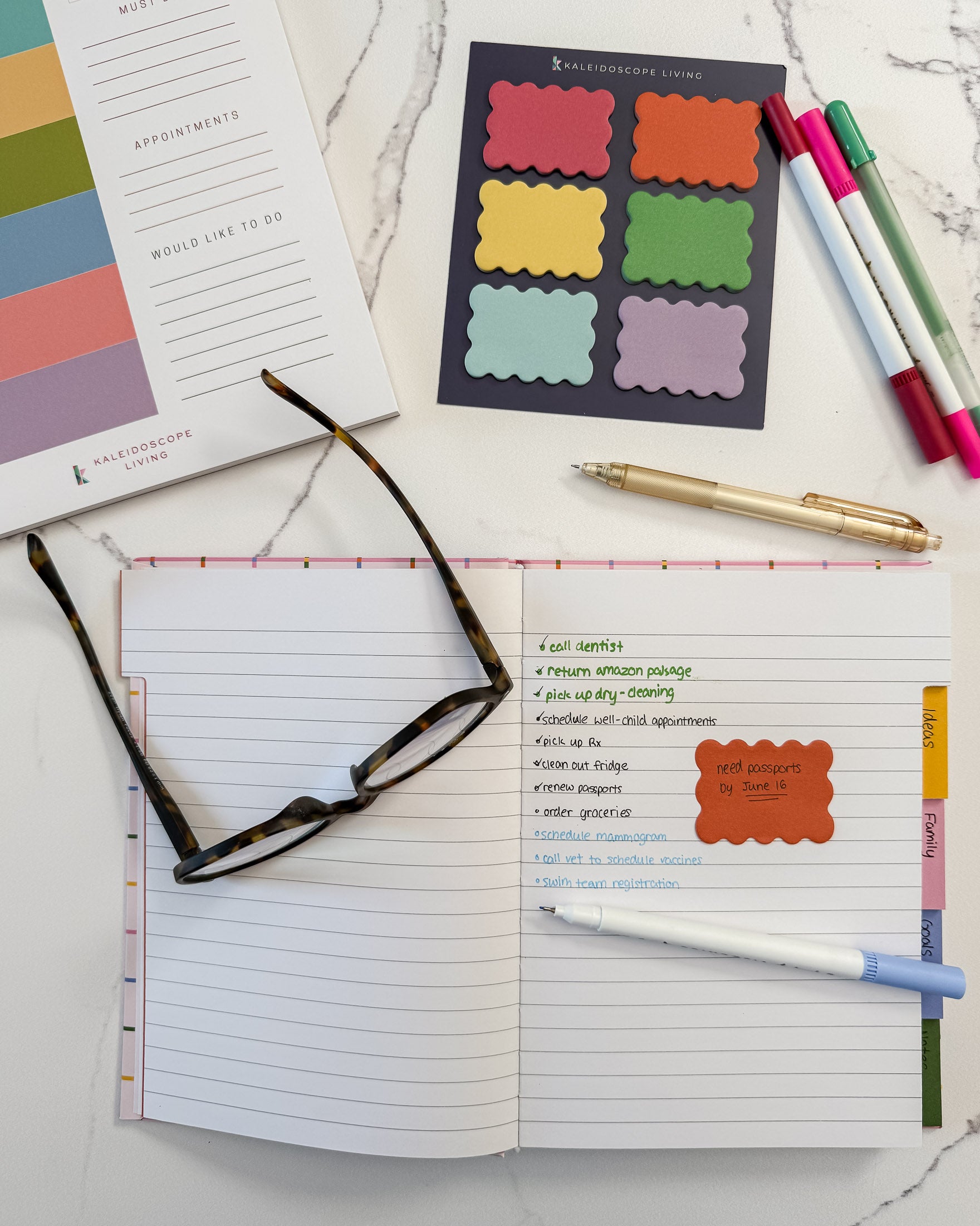 A styled flat-lay of stationery items on a white marble surface, including an open lined notepad with a checklist, a pair of tortoiseshell glasses, colorful scalloped sticky notes, pastel markers, a gold pen, and a notepad from Kaleidoscope Living.