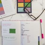 A styled flat-lay of a tabbed notebook with lined pages showing handwritten lists and reminders set on a white marble background with colorful scalloped sticky notes, eyeglasses, and a pink marker nearby.