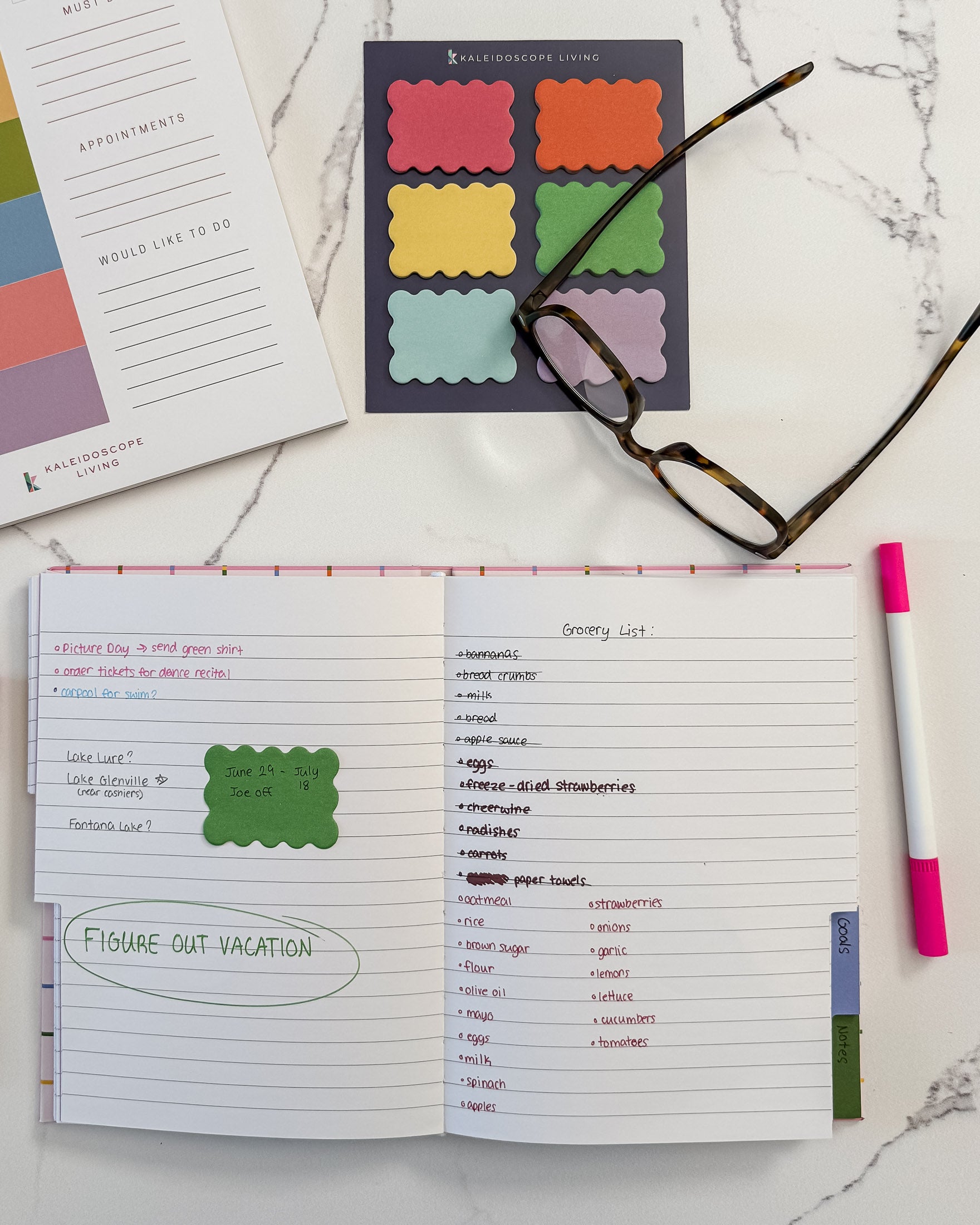 A styled flat-lay of a tabbed notebook with lined pages showing handwritten lists and reminders set on a white marble background with colorful scalloped sticky notes, eyeglasses, and a pink marker nearby.