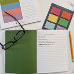 An open notebook with a green page divider and lined pages lies on a white marble surface, styled with a pair of eyeglasses, a yellow pencil, colorful scalloped sticky notes, and a weekly planner notepad.