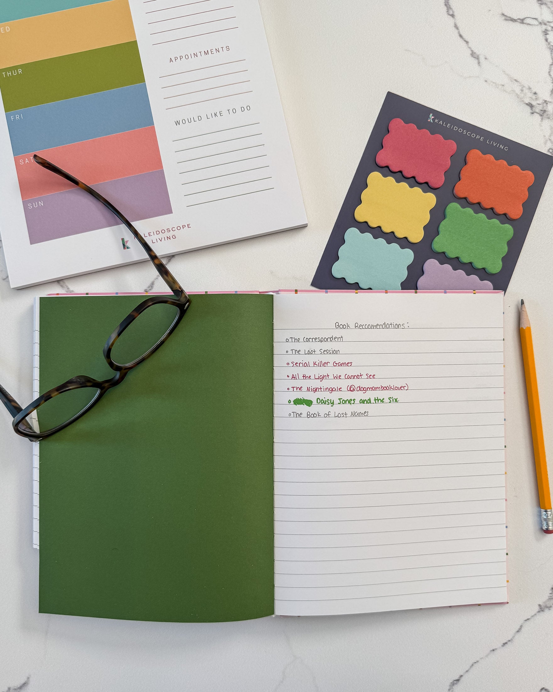 An open notebook with a green page divider and lined pages lies on a white marble surface, styled with a pair of eyeglasses, a yellow pencil, colorful scalloped sticky notes, and a weekly planner notepad.