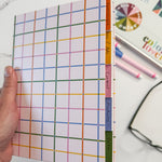 A hand holding a hardcover notebook with a colorful rainbow grid pattern and give tabbed sections labeled 'Contents', 'Products', 'Plan', and 'Notes'. A marble surface, eye glasses, and markers are visible in the background.