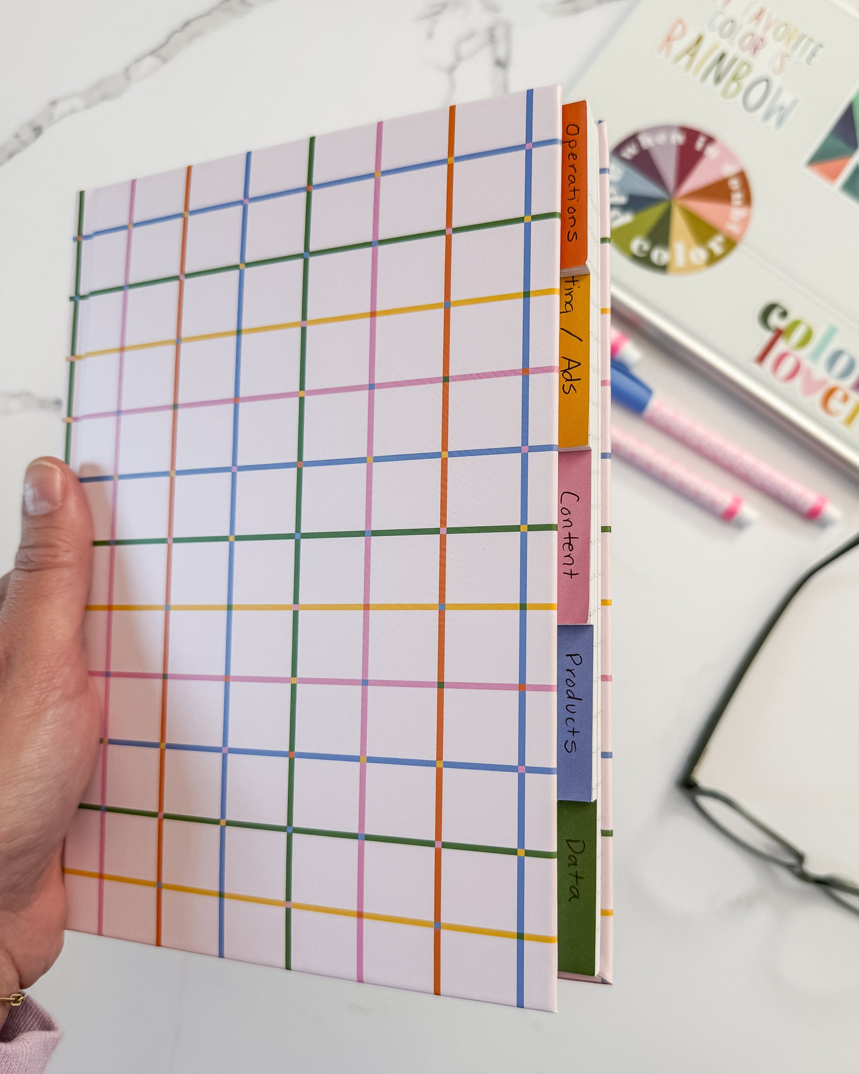A hand holding a hardcover notebook with a colorful rainbow grid pattern and give tabbed sections labeled 'Contents', 'Products', 'Plan', and 'Notes'. A marble surface, eye glasses, and markers are visible in the background.
