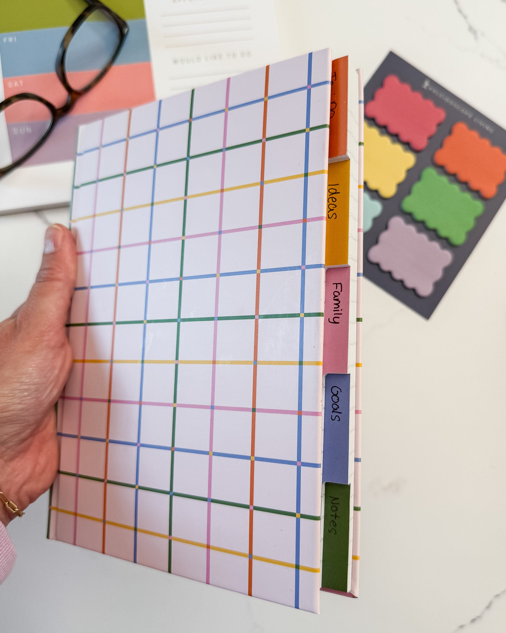 A hand holding a tabbed notebook with a pastel multicolor grid pattern and colorful tab dividers labeled ideas, projects, family, goals, and notes, shown on a white desk with glasses and sticky notes in the background.