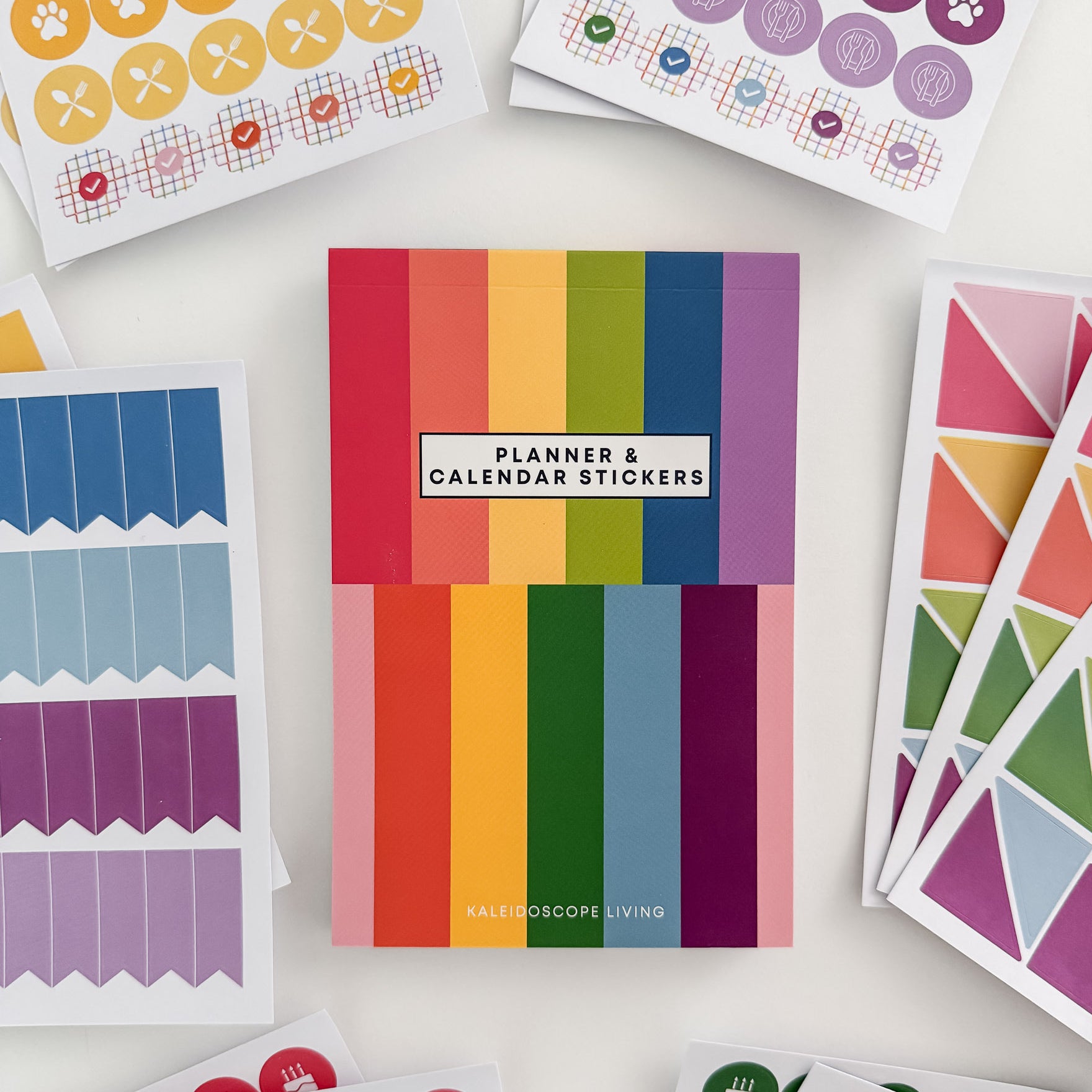 Close up view of Planner and Calendar Stickers top-bound sticker book surrounded by sticker pages featuring a variety of sticker designs in rainbow hues on white surface