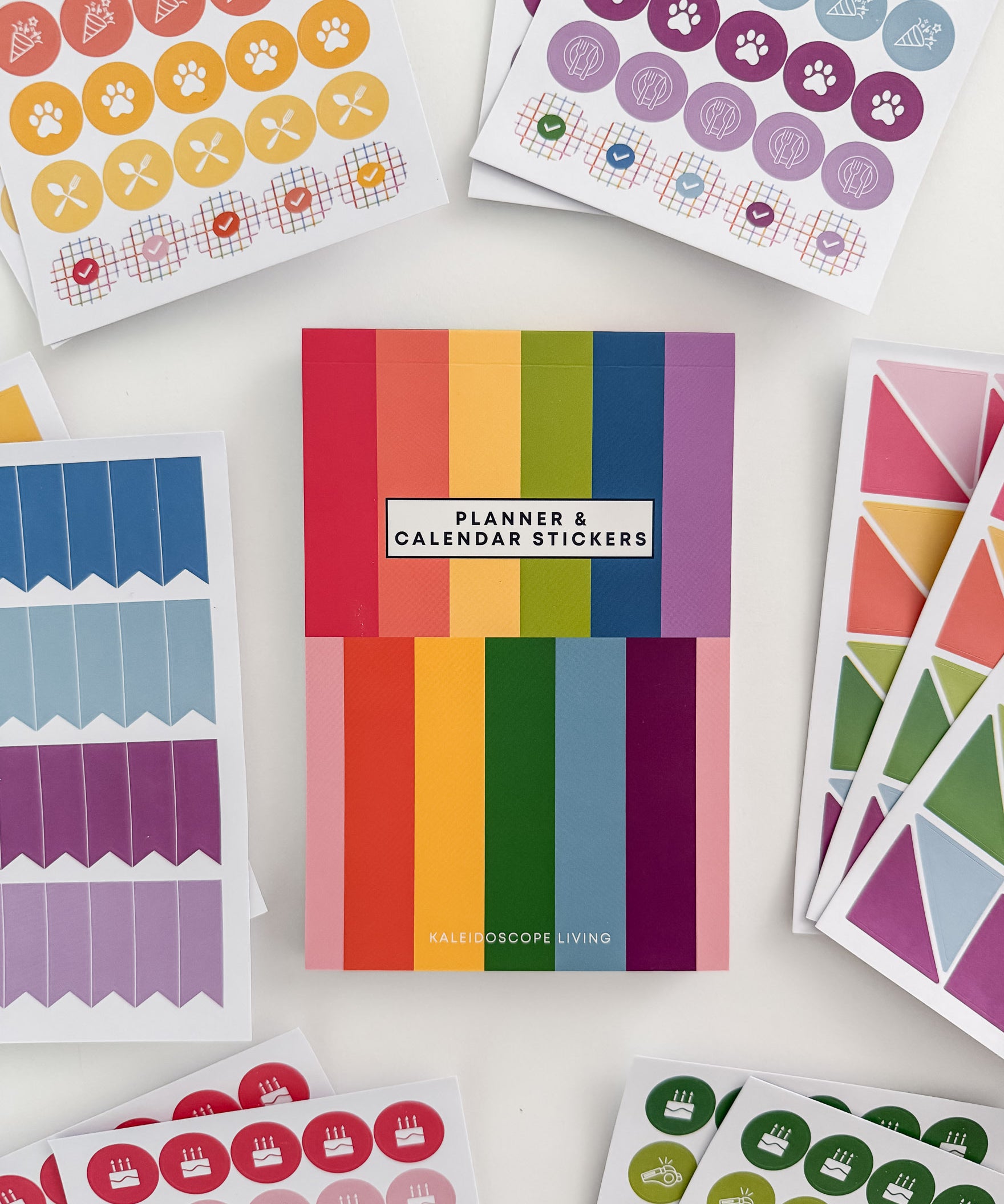 Close up view of Planner and Calendar Stickers top-bound sticker book surrounded by sticker pages featuring a variety of sticker designs in rainbow hues on white surface