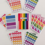 Full view of Planner and Calendar Stickers top-bound sticker book surrounded by sticker pages featuring a variety of sticker designs in rainbow hues on white surface