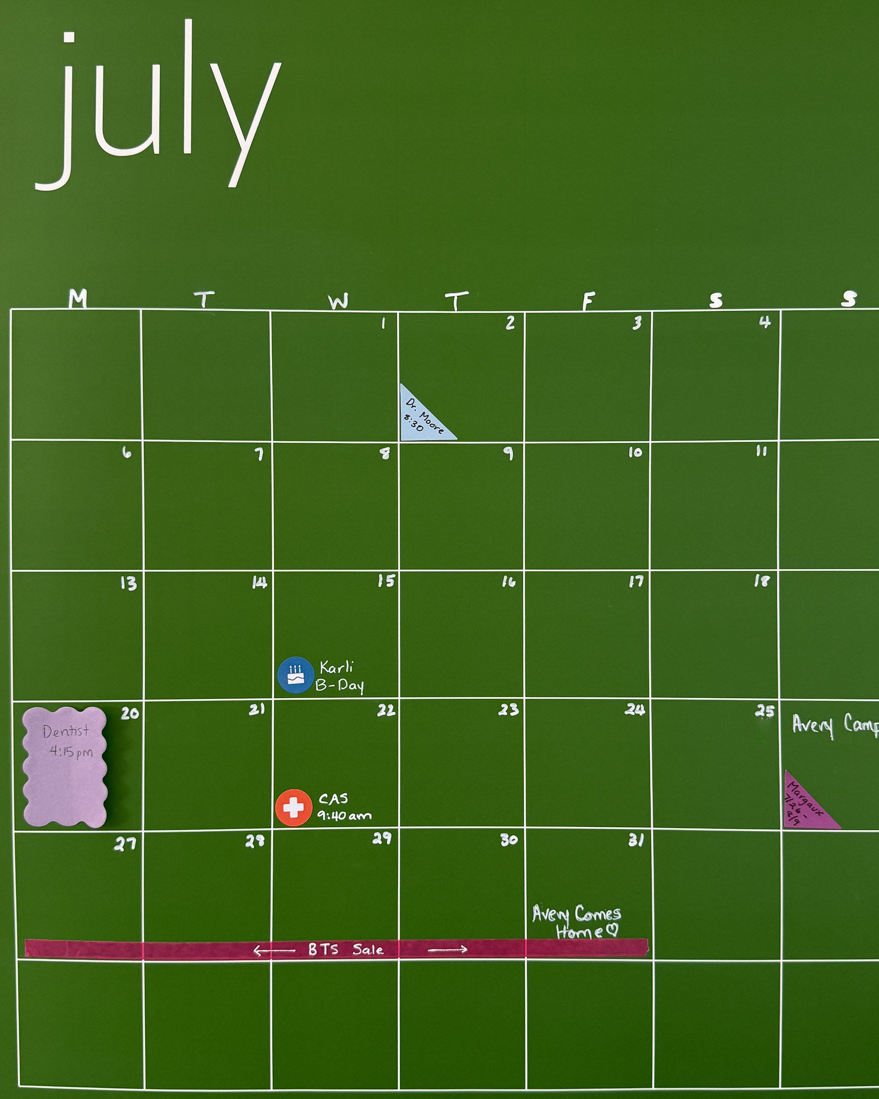 Photo of dark green July wall calendar with colorful planner and calendar stickers, scalloped sticky notes, and washi tape used to mark important appointments and events