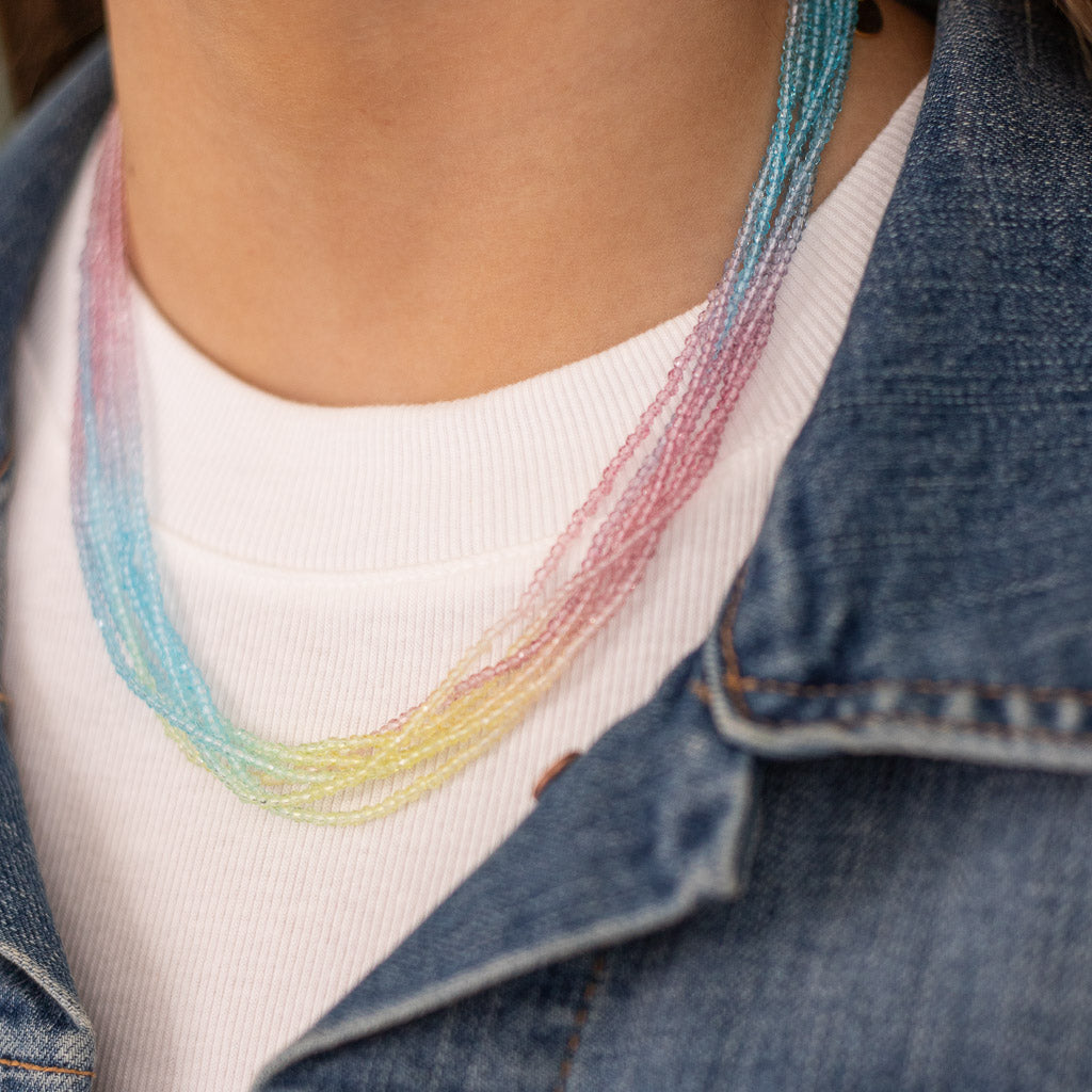 A close-up image of a layered glass seed beaded necklace in shades of pink, yellow, and blue, worn around the neck.