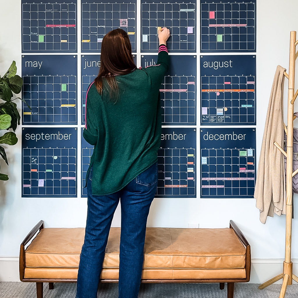 woman adding sticky notes to large reusable wall calendar in navy blue displaying all twelve months