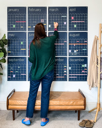 woman adding sticky notes to large reusable wall calendar in navy blue displaying all twelve months