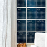 A series of large, navy blue, reusable wall calendar pages arranged in a grid format on a wall, with a tan big sitting on the floor in front of it.