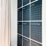 Twelve, large, navy blue, reusable wall calendar pages arranged in a grid format on a white wall.