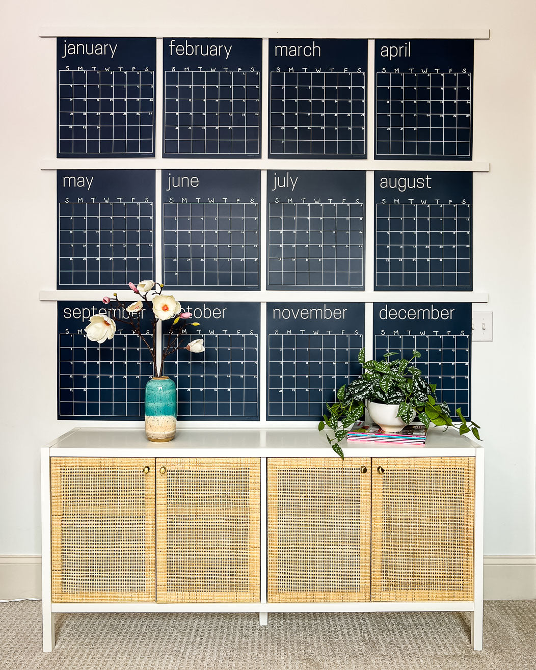 Navy reusable wall calendar from Kaleidoscope Living displayed above a piece of furniture with a flower and plant displayed in front.