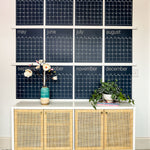 Navy reusable wall calendar from Kaleidoscope Living displayed above a piece of furniture with a flower and plant displayed in front.