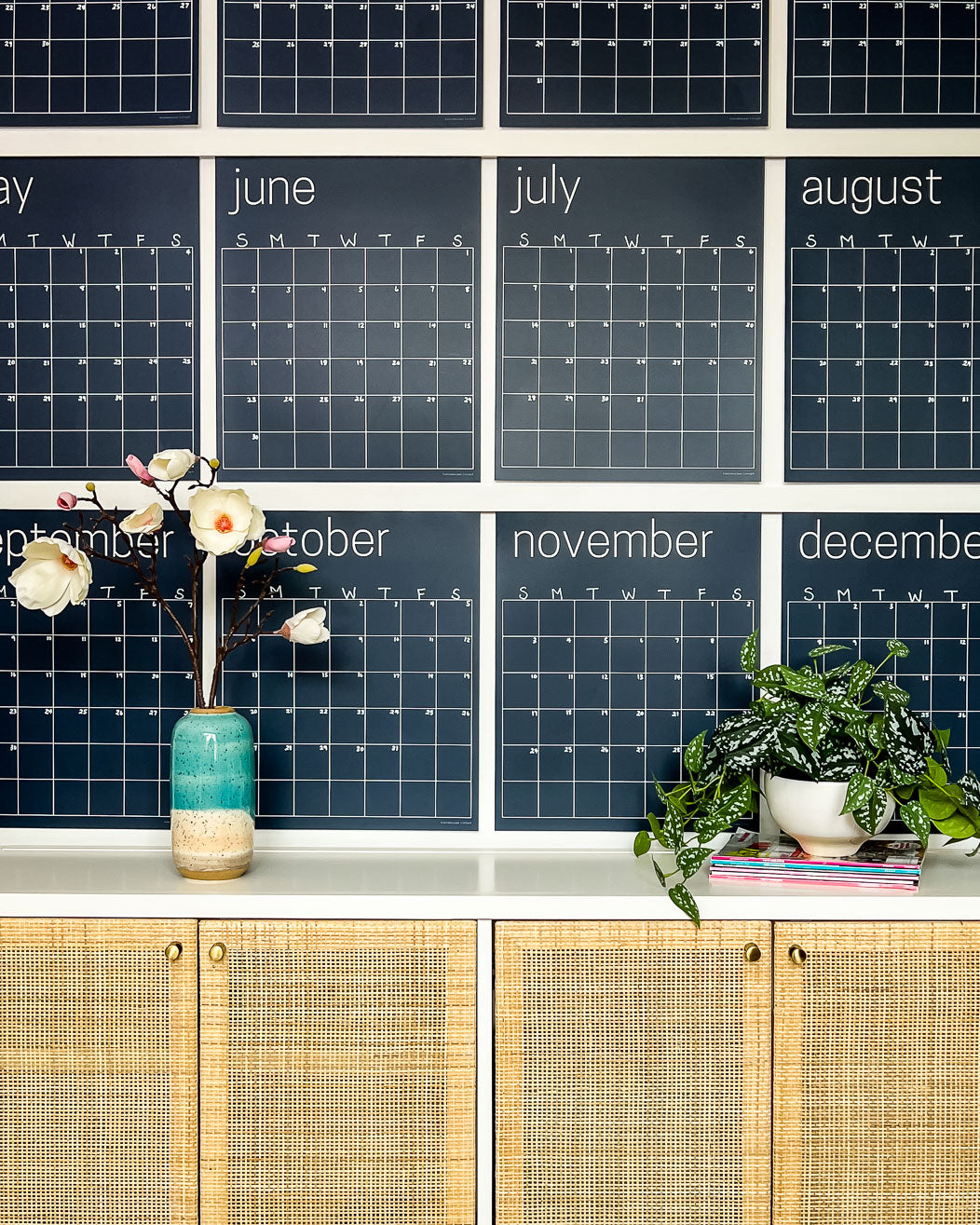 Close up of the navy reusable wall calendar from Kaleidoscope Living displayed above a piece of furniture with a flower and plant displayed in front.