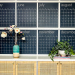 Close up of the navy reusable wall calendar from Kaleidoscope Living displayed above a piece of furniture with a flower and plant displayed in front.