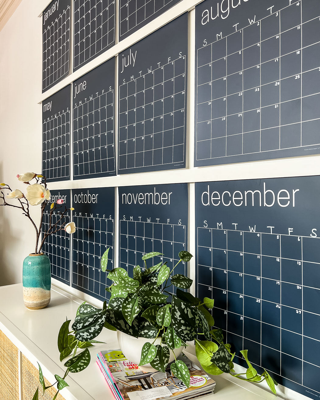 Close up of the navy reusable wall calendar from Kaleidoscope Living displayed above a piece of furniture with a plant and flower sitting in front.