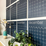 Close up of the navy reusable wall calendar from Kaleidoscope Living displayed above a piece of furniture with a plant and flower sitting in front.
