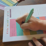 Video of a weekly planner page from Kaleidoscope Living being written on by a woman's hand with notes about with meals, appointments, and to-do lists on a dark wood table.