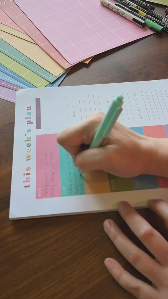 Video of a weekly planner page from Kaleidoscope Living being written on by a woman's hand with notes about with meals, appointments, and to-do lists on a dark wood table.
