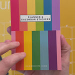 Video showing close up view of flipping through 14 sticker pages of planner and calendar sticker book featuring a variety of sticker designs in rainbow hues with a colorful wall calendar in the background