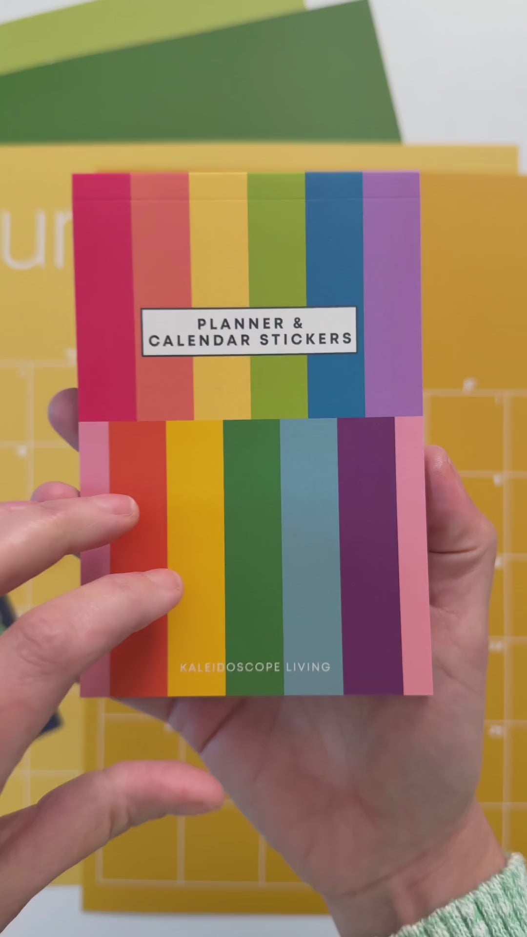 Video showing close up view of flipping through 14 sticker pages of planner and calendar sticker book featuring a variety of sticker designs in rainbow hues with a colorful wall calendar in the background