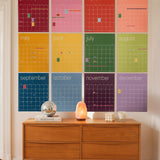 A large 12-month monthly calendar wall art set in a grid of vibrant, solid-colored monthly posters in rainbow color order, displayed on a beige wall above a mid-century modern wooden sideboard with decorative objects.