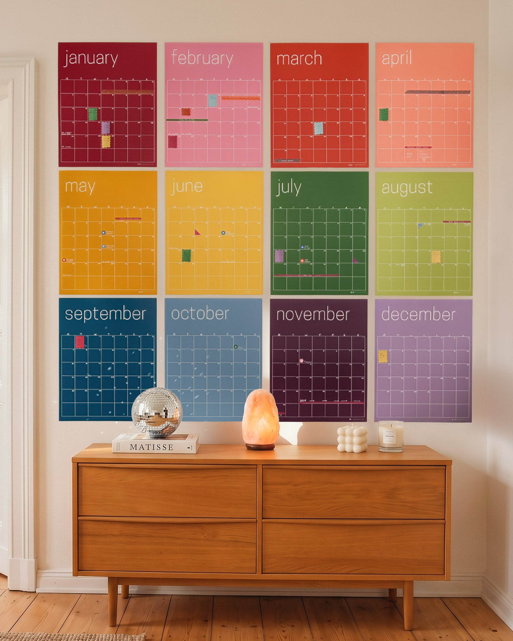 A large 12-month monthly calendar wall art set in a grid of vibrant, solid-colored monthly posters in rainbow color order, displayed on a beige wall above a mid-century modern wooden sideboard with decorative objects.