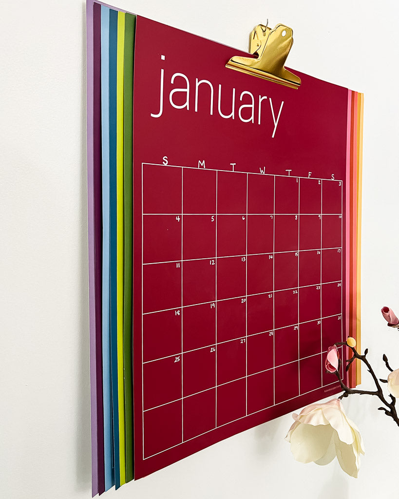 large colorful undated monthly wall calendar hanging with large gold clip on white wall, with all colors visible.