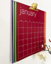 large colorful undated monthly wall calendar hanging with large gold clip on white wall, with all colors visible.